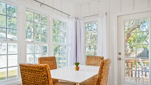 Ein Zimmer mit einem weißen Tisch, Korbstühlen, großen Fenstern und Blick ins Freie.