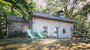 Uma casa de pedra com telhado de telhas, duas janelas e um pequeno alpendre com duas cadeiras de jardim.