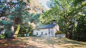 Um edifício de pedra com telhado inclinado, cercado por árvores e uma área gramada com mesa e cadeiras para piquenique.