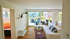 A modern living room with a white sofa, a glass coffee table, and a view of the outdoors through large windows.