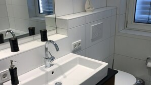 A modern bathroom with a white sink, black faucets, and a mirror above it. There is a toilet to the right and a window with blinds.