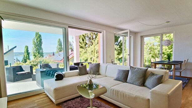 A modern living room with a white sofa, a glass coffee table, and a view of a balcony with outdoor furniture.