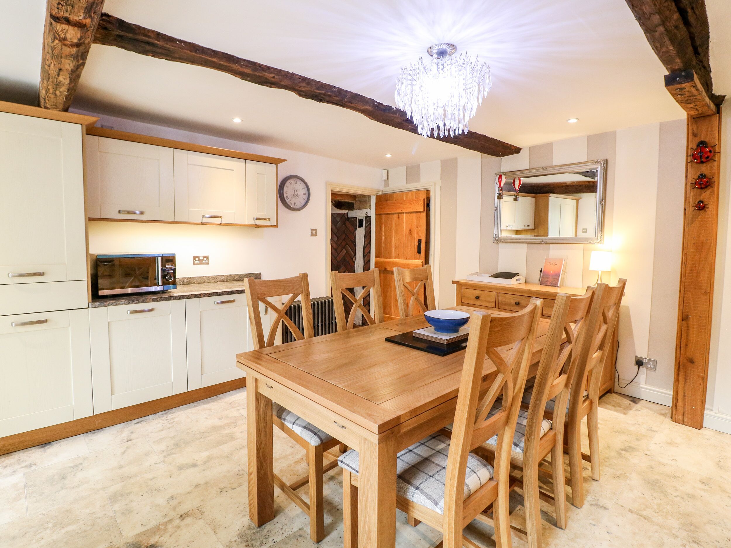 A dining area with a wooden table and chairs, a chandelier, and a kitchen with white cabinets and a microwave.
