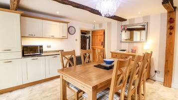 A dining area with a wooden table and chairs, a chandelier, and a kitchen with white cabinets and a microwave.