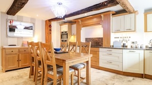 A dining area with a wooden table and chairs, a chandelier, and a kitchen with white cabinets and a marble backsplash. - Christmas Cottage (Alfreton)