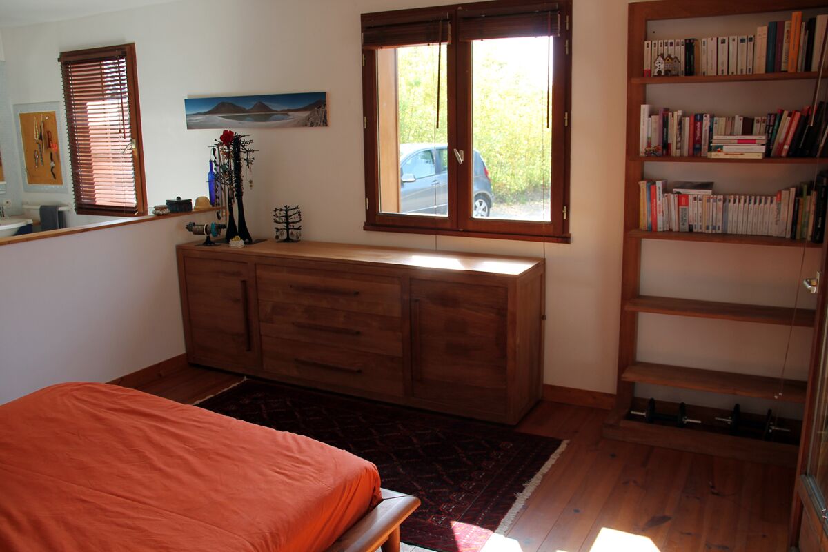 A bedroom with a bed, a wooden dresser, a bookshelf, and a window with a view of a car and greenery outside.