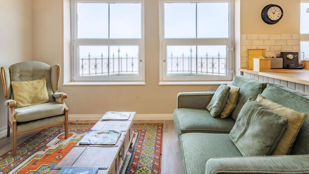 A living room with a green sofa, a wooden chair, a coffee table with books, and a colorful rug. There are two windows with a view of a fence, and a kitchen area with a clock and appliances.