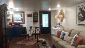 Sala de estar con sofá, mesa de centro y sillas. Se observa una zona de cocina con refrigerador, microondas y pileta. En el fondo, se ve una puerta con ventana.