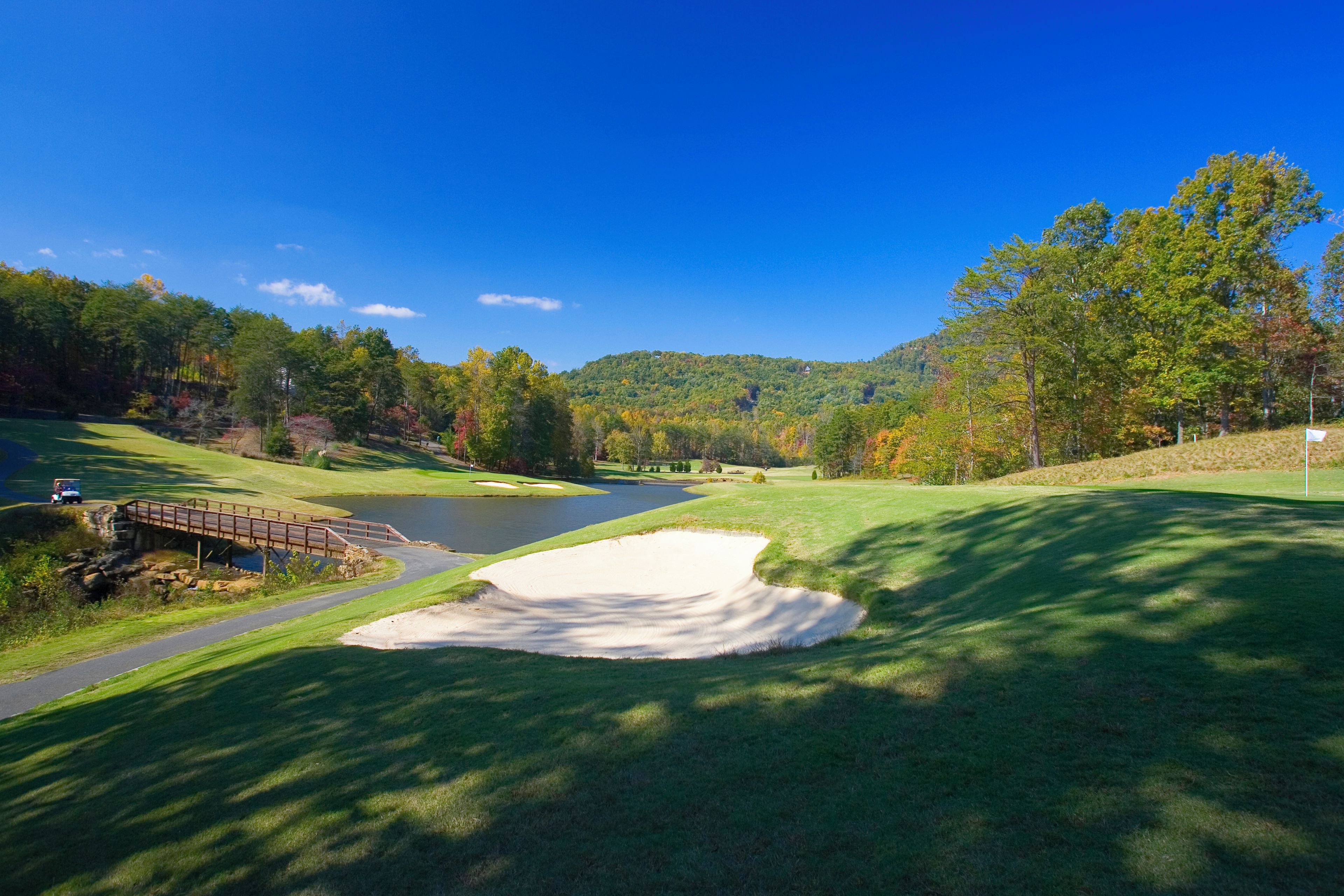 A golf course with a pond, a bridge, and a forested hill in the background.