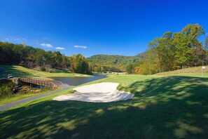 A golf course with a pond, a bridge, and a forested hill in the background.