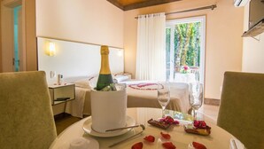 A hotel room with a dining table set for two, a minibar, a bed with a white canopy, and a view of greenery through the window. - Pousada Recanto do Tirol (Gramado)