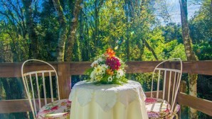 A table with a floral centerpiece, two white chairs, and a wooden deck with a forest backdrop. - Pousada Recanto do Tirol (Gramado)