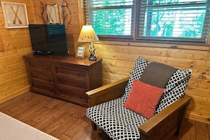 A wooden cabin interior with a wooden dresser, a television, a lamp, and a chair with patterned cushions.
