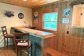 A wooden bar area with two chairs, a wooden counter, and a window with a view of greenery.