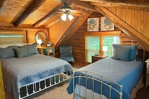 A cozy attic bedroom with two beds, wooden walls, a ceiling fan, and artwork.