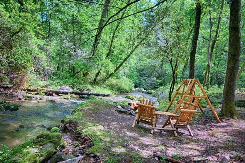A forest with a stream, a wooden swing set, and two chairs.