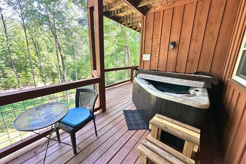 Eine Holzterrasse mit Whirlpool, einem Tisch und einem Stuhl, mit Blick auf einen Wald.