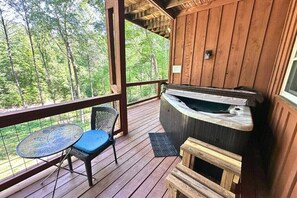 Un terrazzo in legno con una vasca idromassaggio, un tavolo e una sedia, e vista su una foresta.