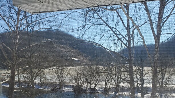A view from a covered deck overlooking a snowy landscape with bare trees and a distant hill. - Cozy Riverfront Cabin (Mendota)