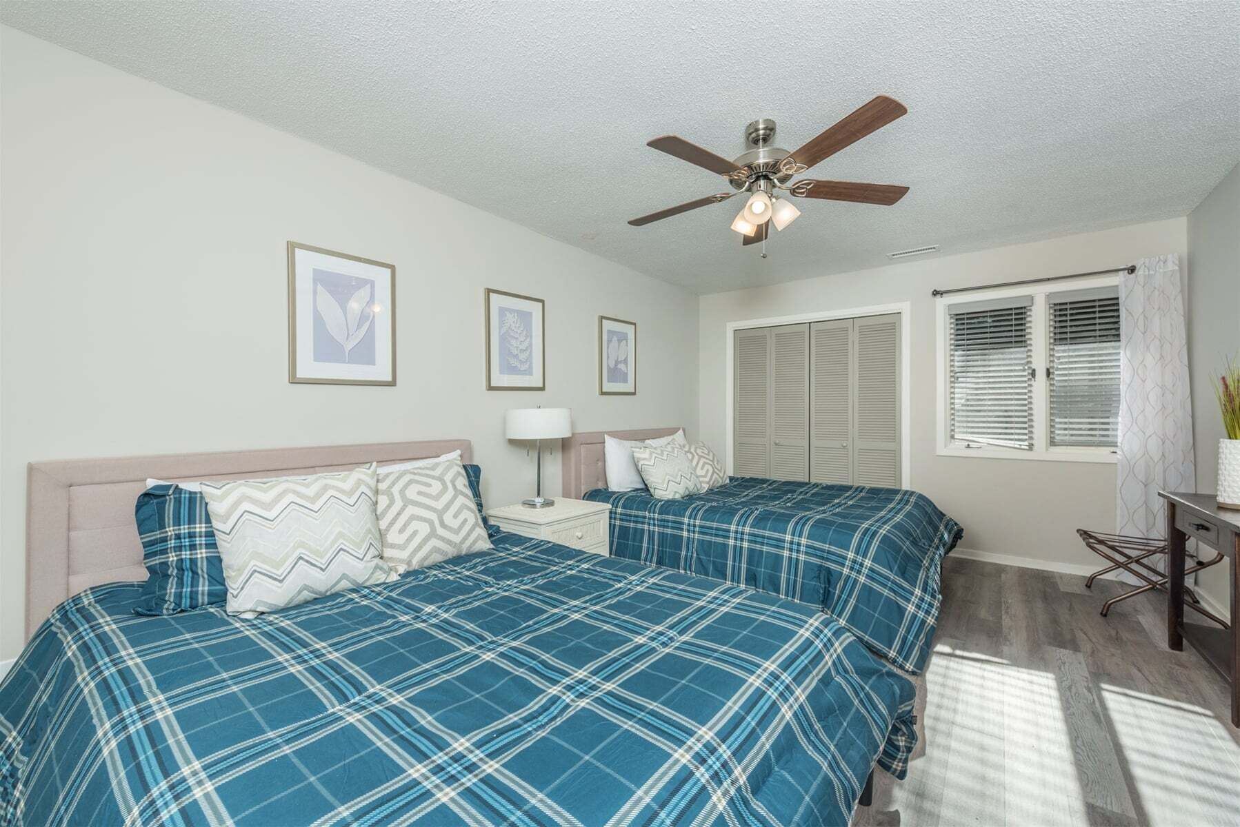 A bedroom with two beds, a ceiling fan, framed artwork, a nightstand, a lamp, a window with blinds, and a small table.
