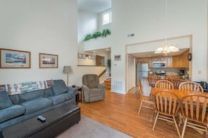 A spacious living room with a sofa, armchair, and dining table. A staircase leads to a kitchen area.