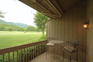 A wooden deck with a table and chairs, overlooking a grassy area with trees and hills.