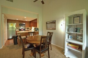 A dining area with a round table and chairs, a kitchen in the background, and a bookshelf with various items.