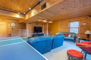 A room with a ping pong table, a sofa, a TV, and a window.