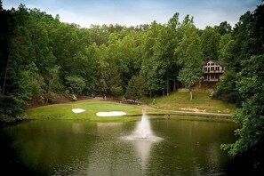 A golf course with a pond, a fountain, and a house in the background.