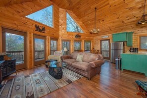 A wooden cabin interior with a living room, kitchen, and large windows.
