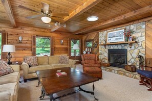A log cabin interior with a stone fireplace, wooden furniture, and a ceiling fan.