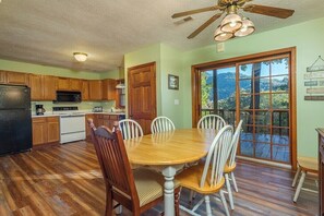 A kitchen with wooden cabinets, a dining table with chairs, a ceiling fan, and a large window with a view.