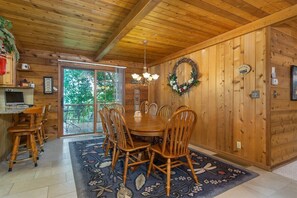 A wooden interior with a dining table, chairs, a chandelier, and a view through a glass door.