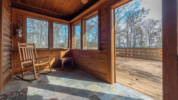 A log cabin interior with a rocking chair, a small table, and large windows offering a view of the forest.