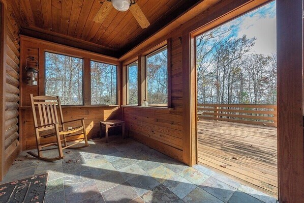A log cabin interior with a rocking chair, a small table, and large windows offering a view of the forest.