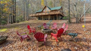 Une cabane dotée d’une terrasse, entourée de chaises Adirondack rouges, d’un foyer et bordée par une forêt.