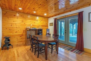 A wooden interior with a dining table, chairs, a stove, and a view through glass doors.