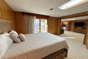 A bedroom with a large bed, wooden walls, a sliding glass door to a balcony, and a kitchenette area.
