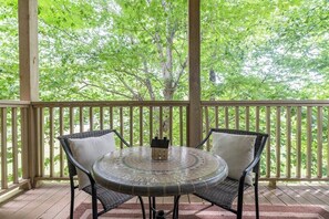 A covered outdoor area with a round table and two chairs, a small house decoration, and a view of green trees.