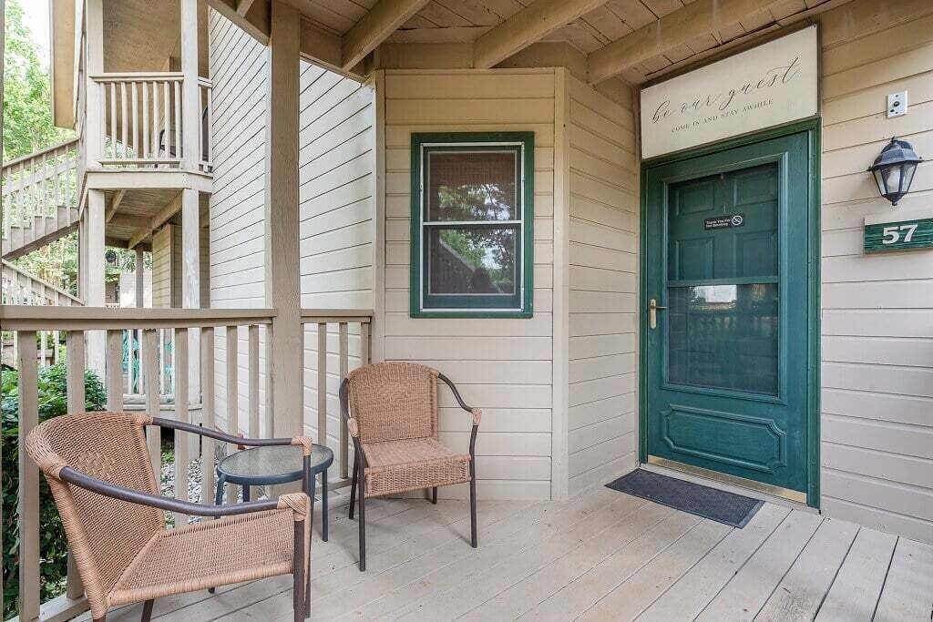 A porch with wicker chairs and a table, a green door with a sign, and a wooden staircase.