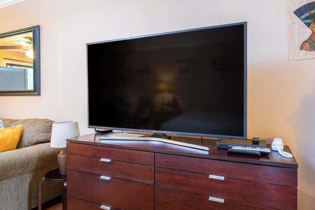A hotel room with a large flat-screen TV on a wooden dresser, a lamp, a remote control, and a telephone.
