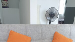 A grey sofa with two orange cushions and a standing fan in the background. - T1 apartment in the city center of Grand-Baie 2 minutes from the beach (Grand Baie)