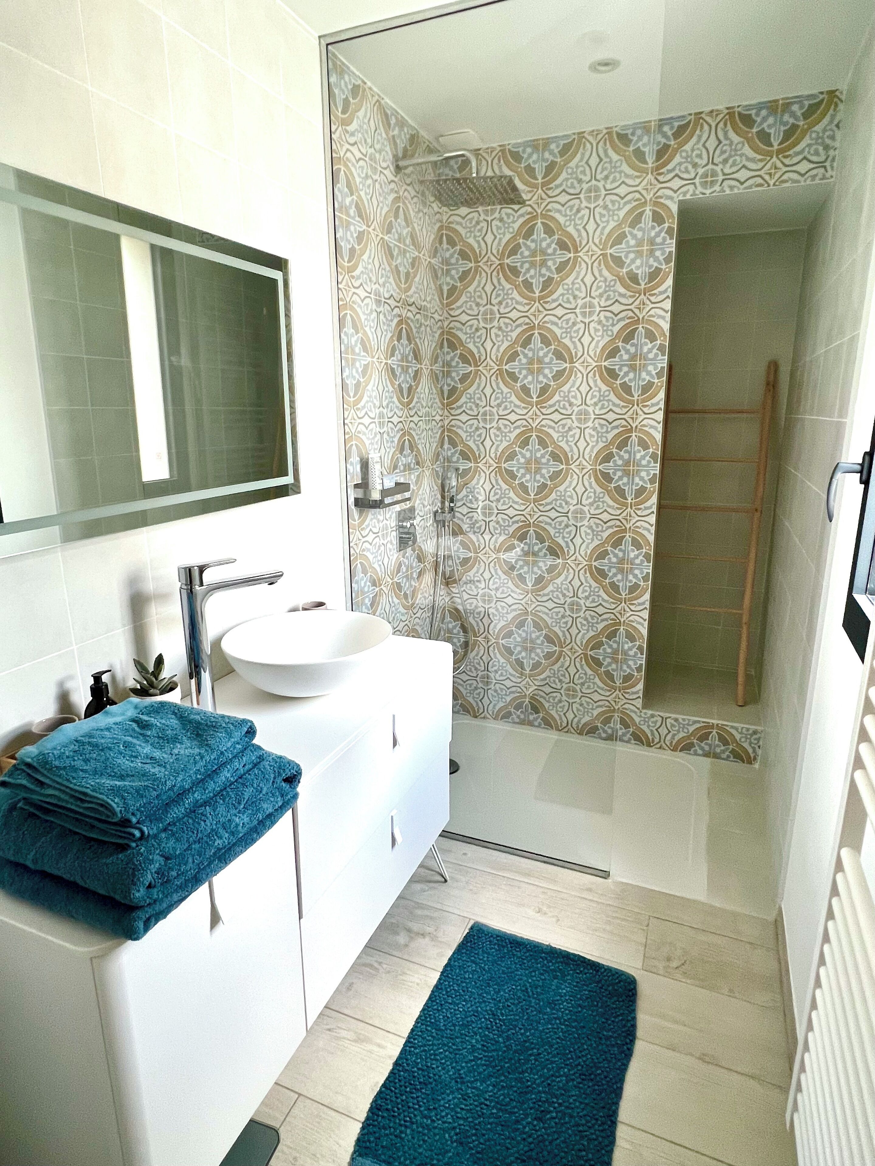 Een badkamer met een witte wastafel, spiegel en blauwe handdoeken. Er zijn betegelde vloeren en een douchegedeelte met een trap.