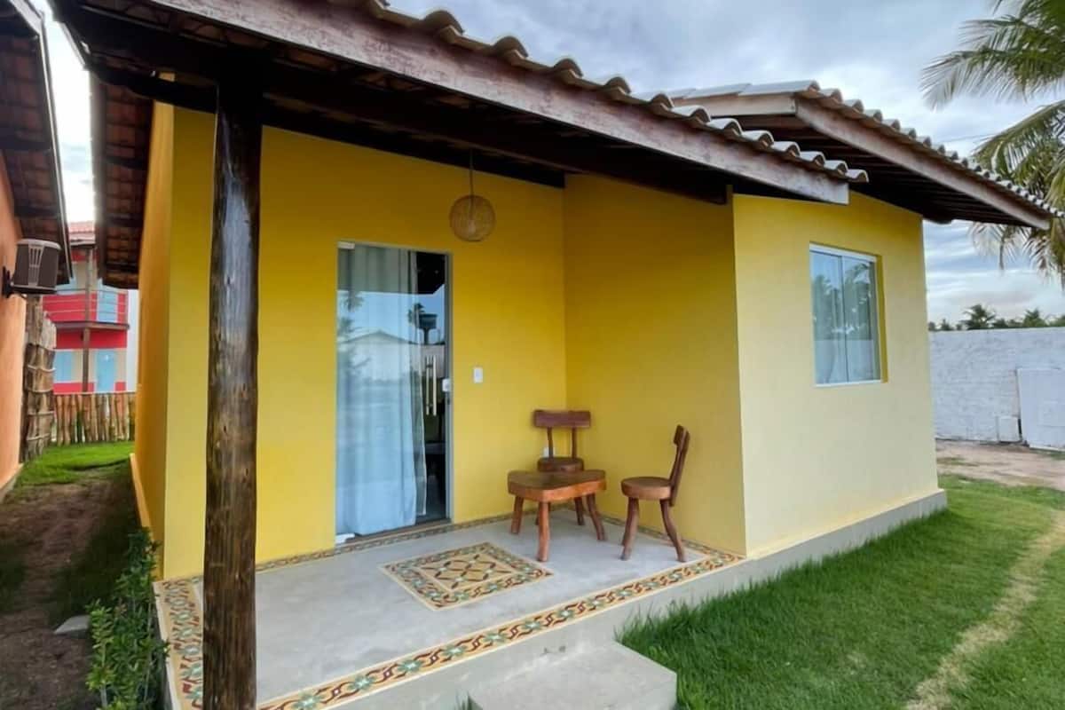 A small yellow building with a tiled roof, a porch with two wooden chairs, and a small table.