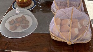 Preparación para un desayuno al aire libre con una mesa llena de comida y bebidas, rodeada de sillas de madera y un área techada con vista a una piscina y palmeras.