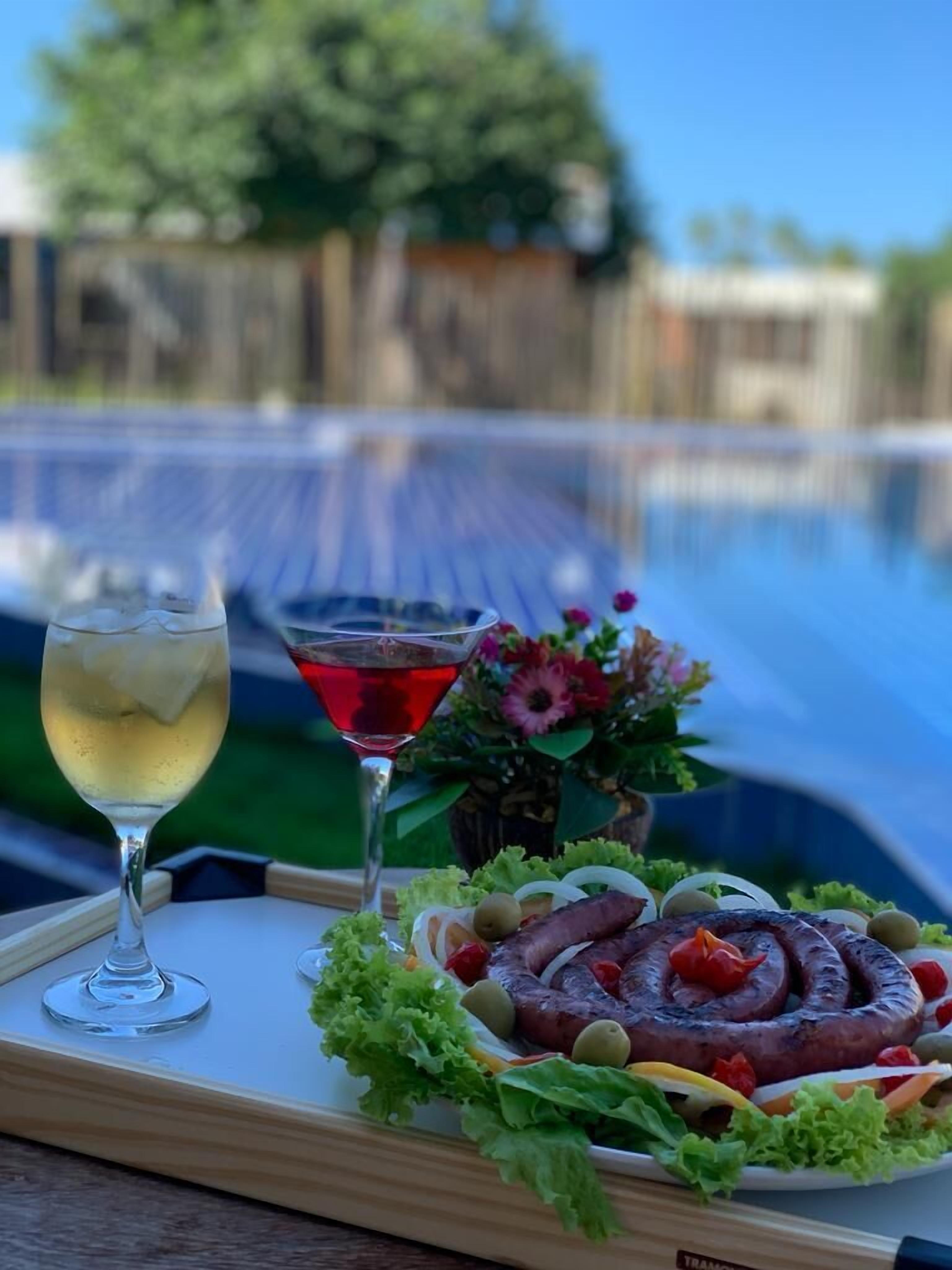 A tray with a glass of white wine, a red cocktail, and a plate of food including sausages, olives, and vegetables.