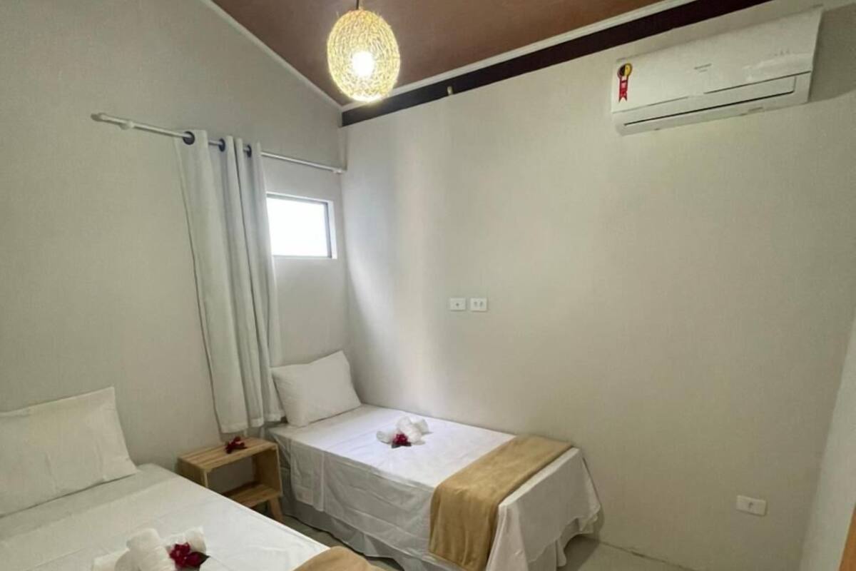A small, clean room with two beds, a window with curtains, an air conditioning unit, and a hanging light fixture.