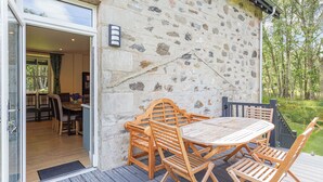 A wooden deck with a round table and chairs, a stone wall, and a door leading to an interior space with a dining area.