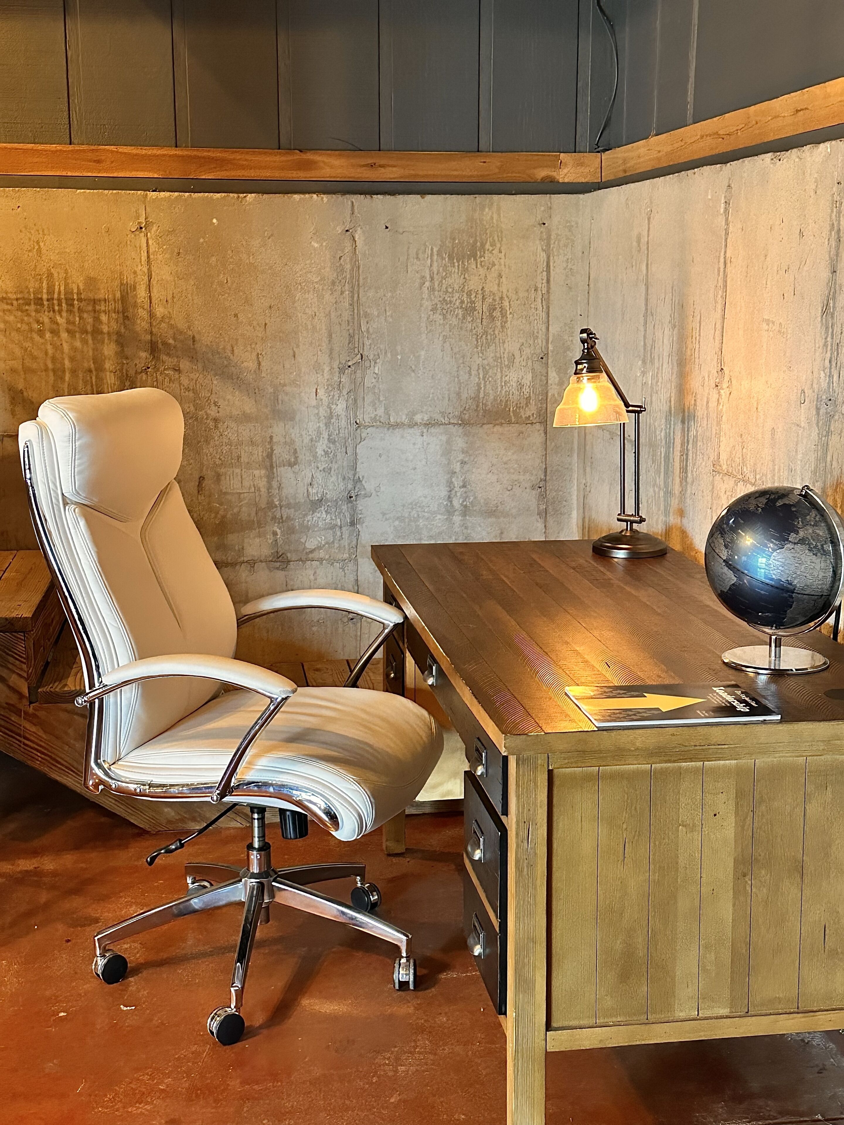 Un bureau en bois avec un globe terrestre et une lampe, un fauteuil en cuir et un mur en béton.