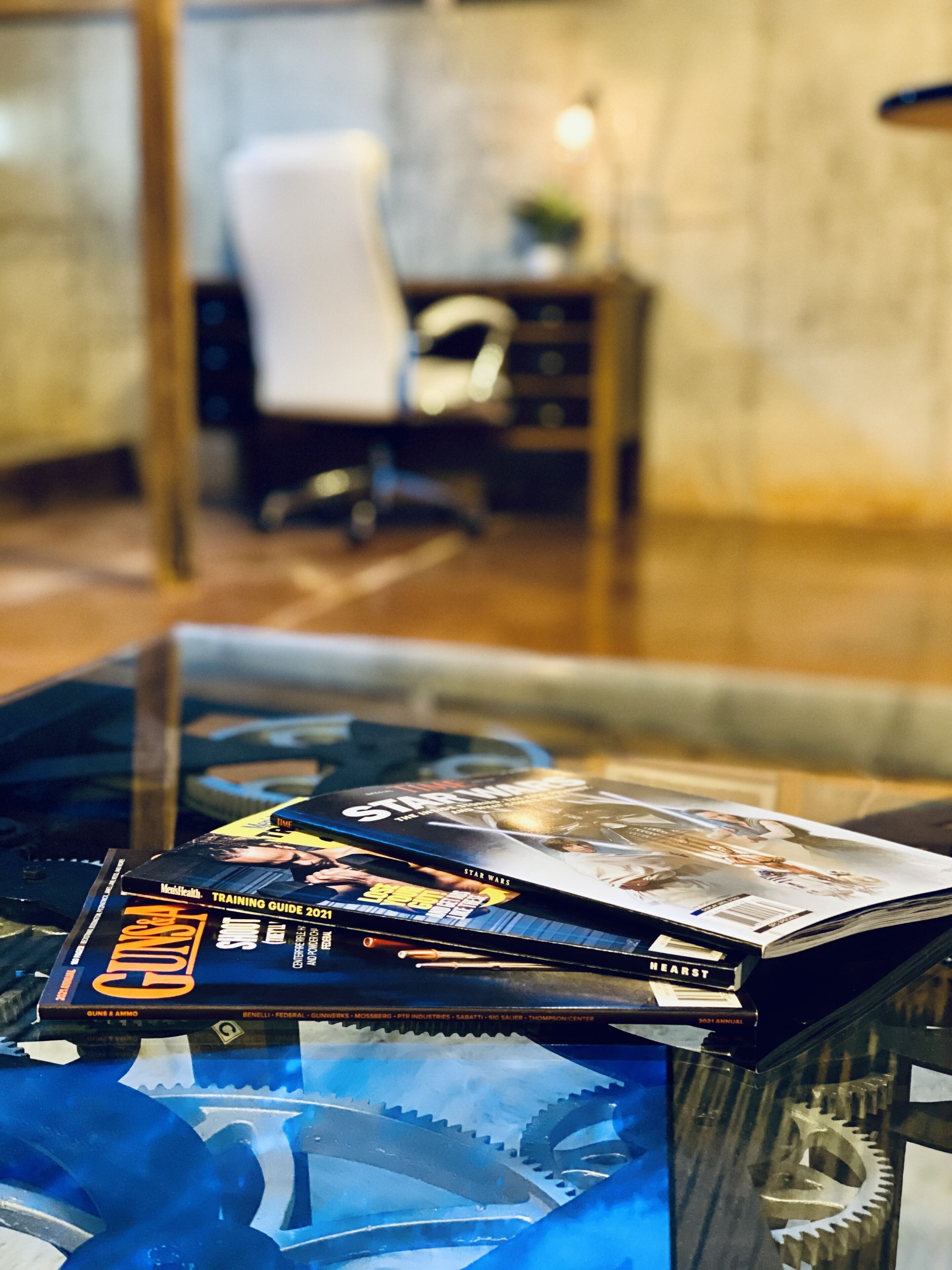 A glass table with magazines and books, a chair, and a desk in the background.